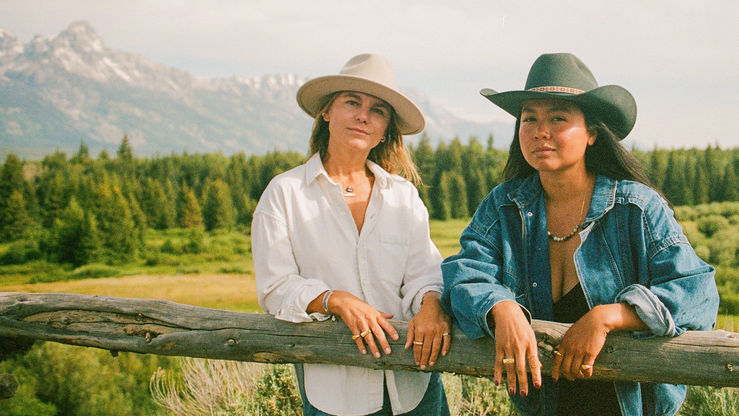 Two women posing outdoors in a scenic mountain landscape, leaning on a rustic wooden fence. One wears a beige wide-brim hat and a white button-down shirt, while the other wears a black cowboy hat and a blue denim shirt. They both are wearing gold jewelry.