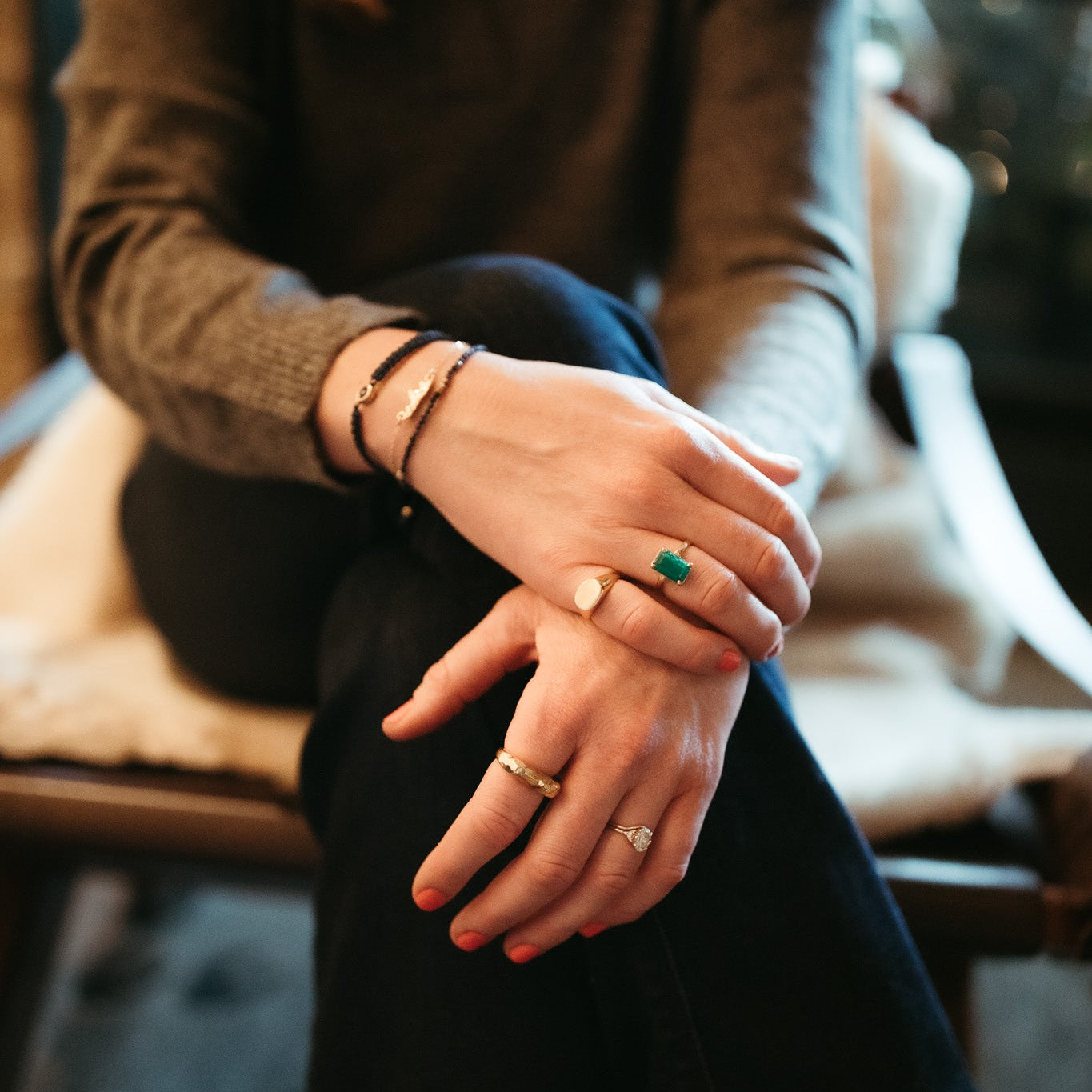 Person sitting with hands clasped, wearing multiple rings and bracelets and gold signet ring on pinky finger, minimalist jewelry style