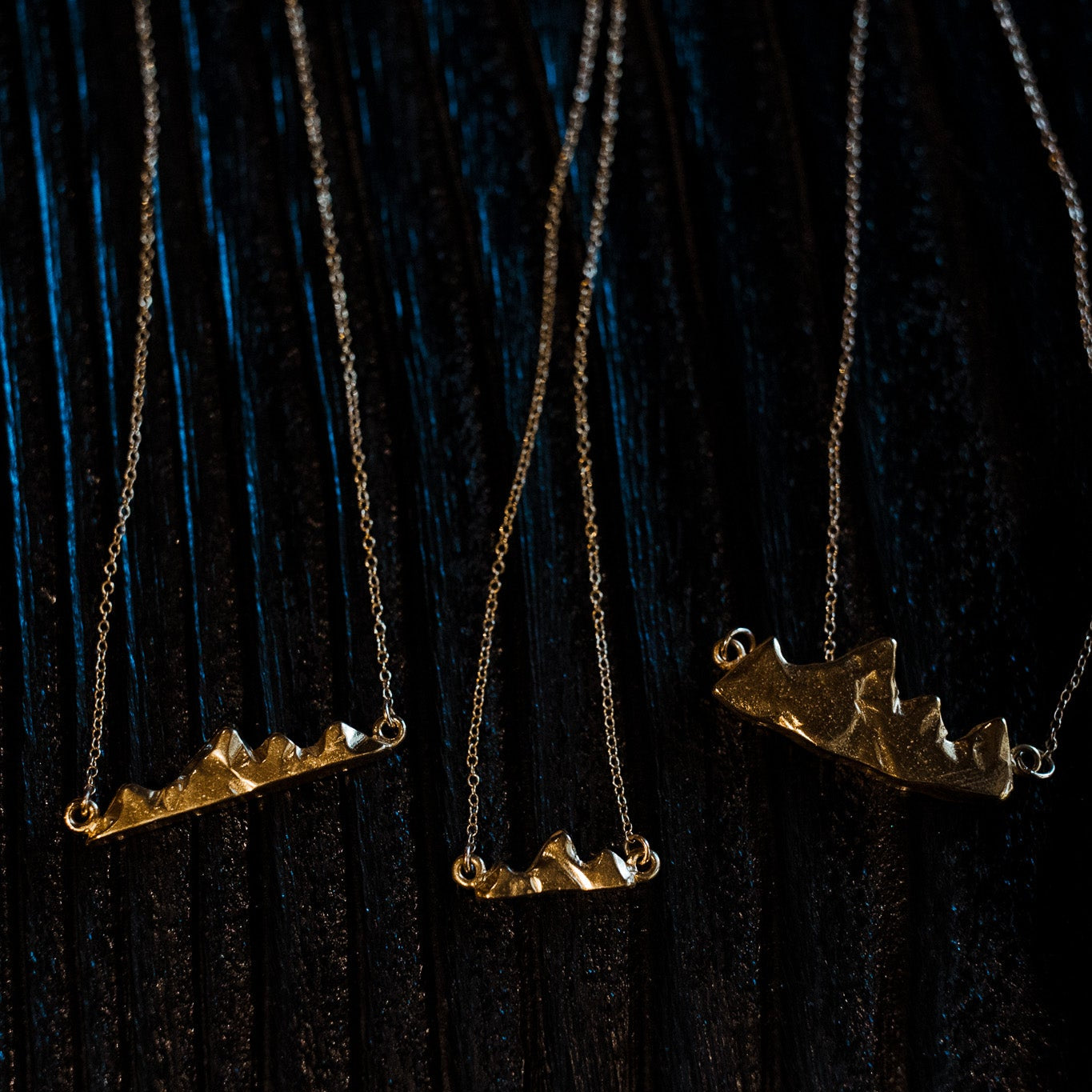 Close-up of delicate Teton I, II, and III pendant showing miniature mountain detail on a black wood surface
