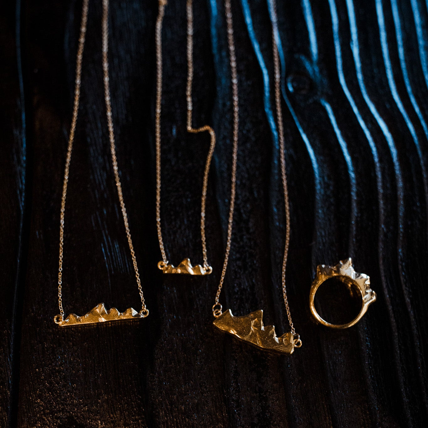 Close-up of Teton I, II, and III pendants and the Teton ring showing refined mountain silhouette on black wood surface