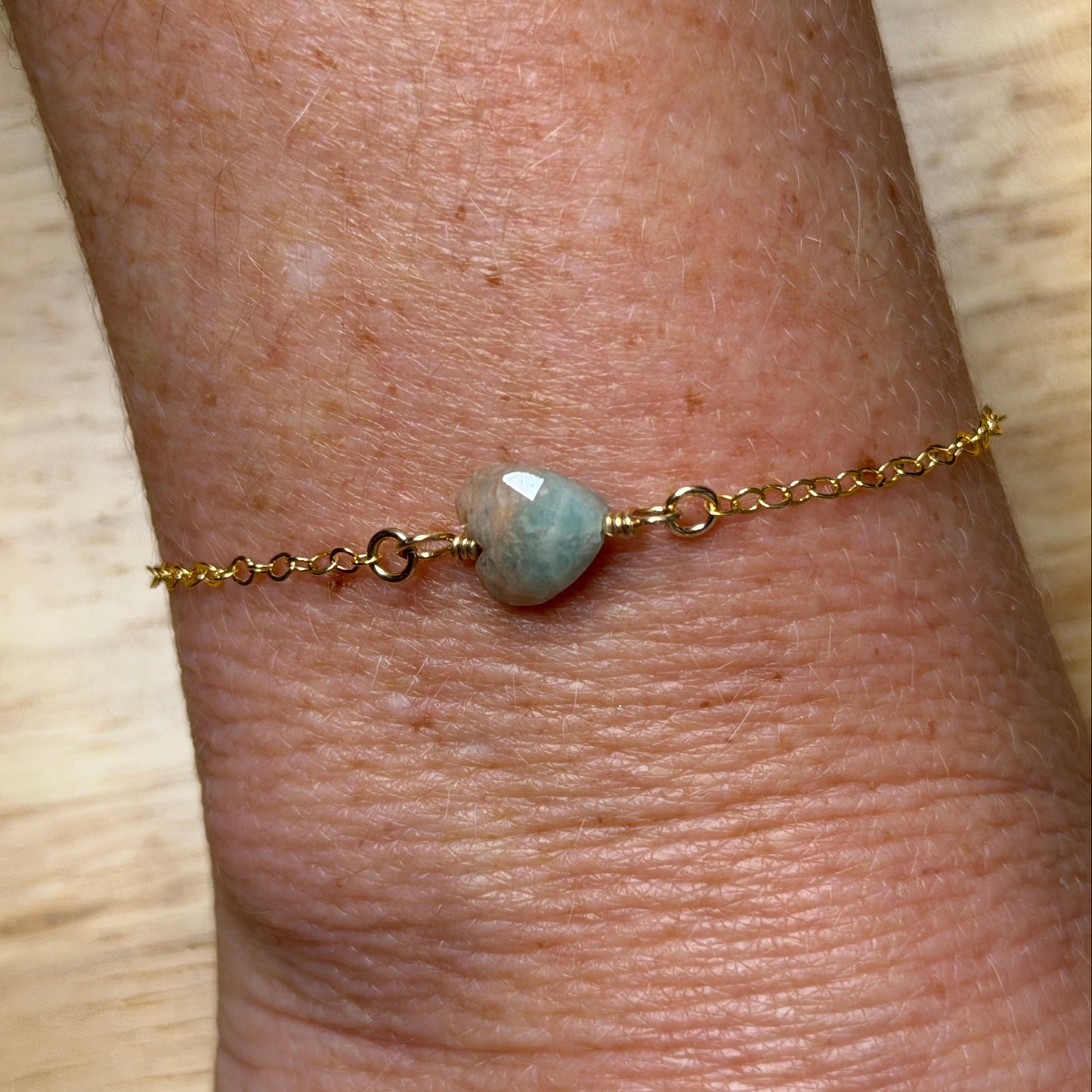 Gold filled wire wrapped amazonite heart bracelet on a gold filled chain on a wrist against a wooden surface