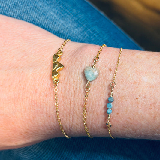 Close-up of a wrist wearing three gold bracelets, one is the Teton Bracelet with different designs on a blue denim background.
